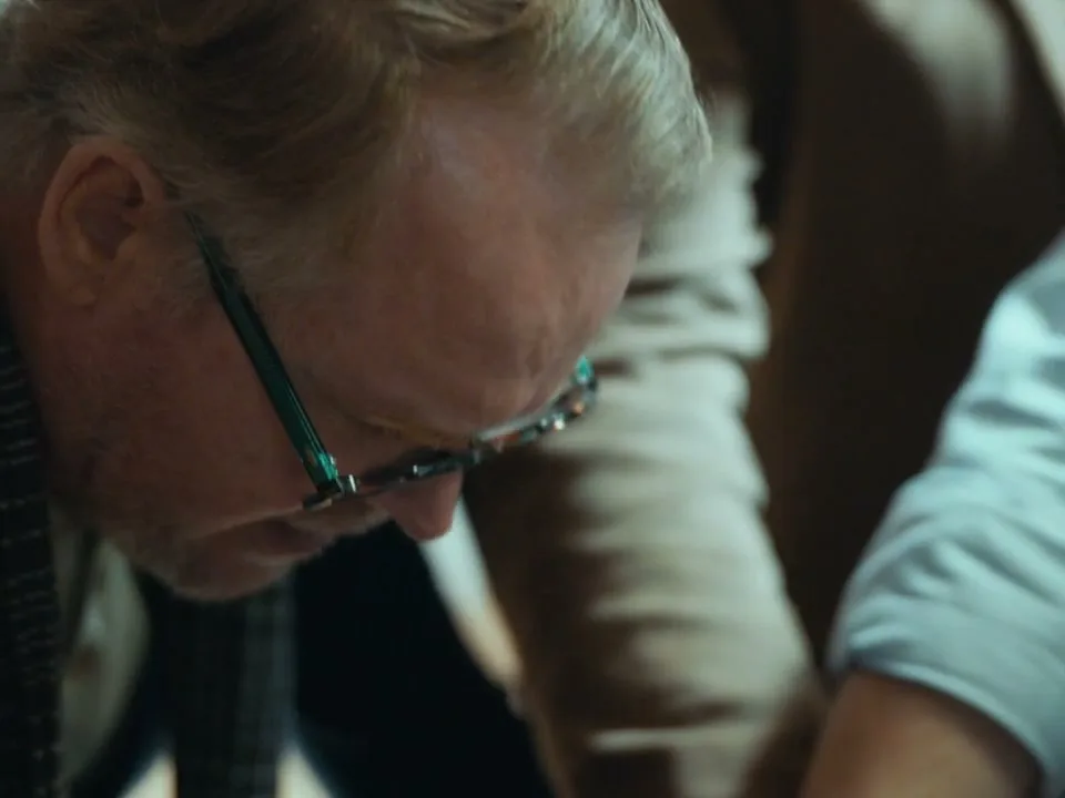 A man with blonde hair and glasses looks down, his face partially obscured, in a dimly lit interior setting.