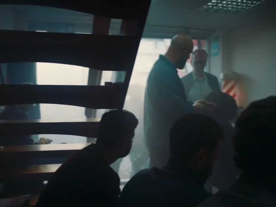 A barber in a blue shirt cuts a man's hair in a barbershop, while other men wait, and a wall of headshots is visible.