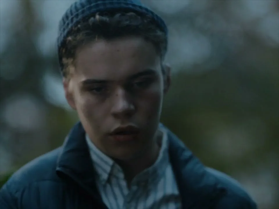 A young man with a blue beanie and jacket hugs an older person, then looks directly at the camera with a somber expression in an outdoor setting.