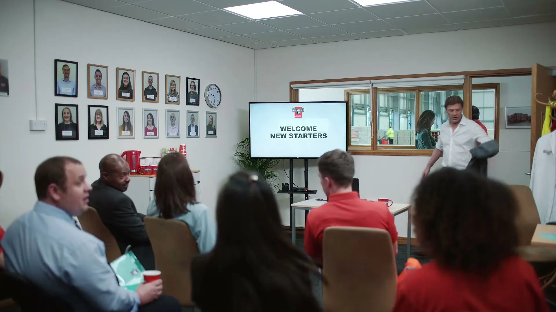 A man in a white shirt walks into a meeting room where a group of people are seated, facing a screen that reads "WELCOME NEW STARTERS."