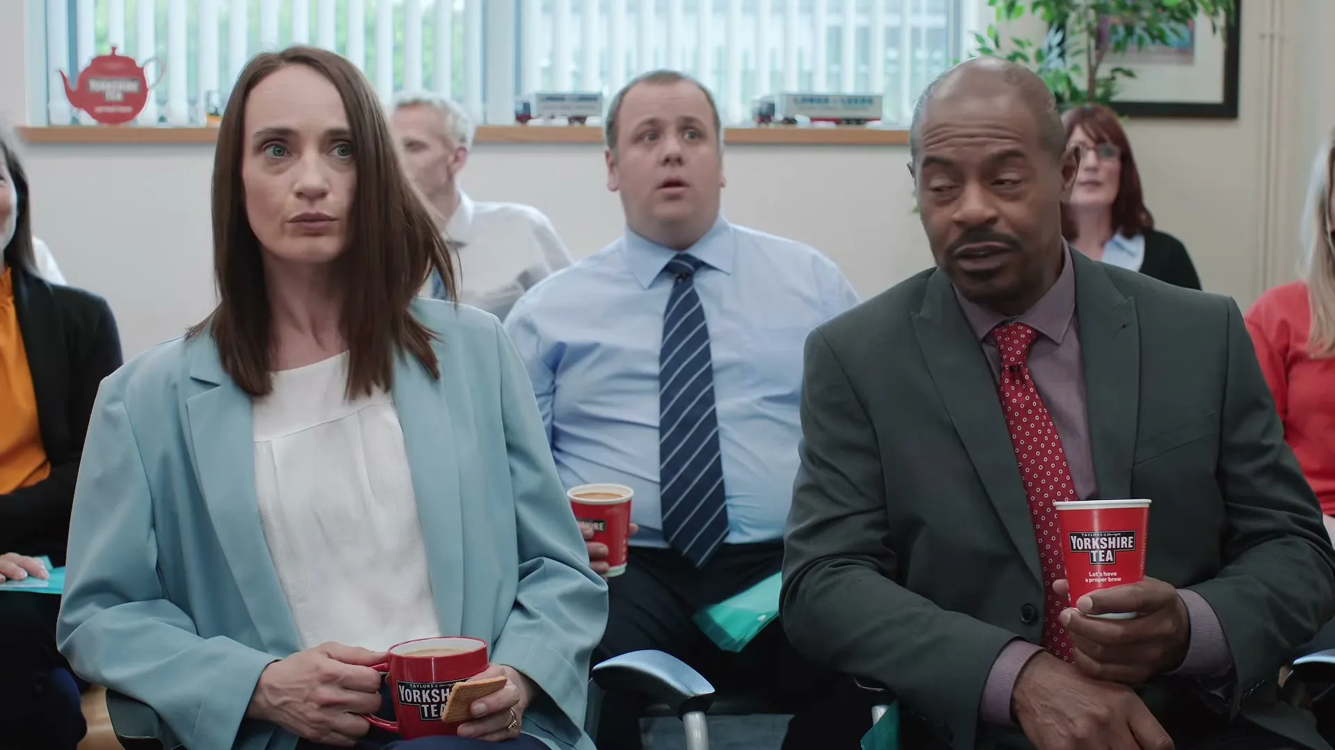 A diverse group of office workers sit in a meeting room, holding cups of Yorkshire Tea, looking around with confused expressions.