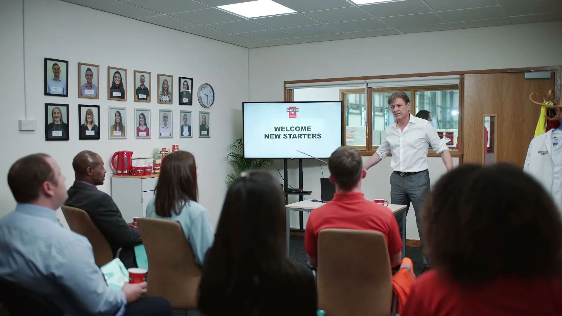 A man in a white shirt presents to a group of new employees in an office meeting room with a screen displaying "WELCOME NEW STARTERS".