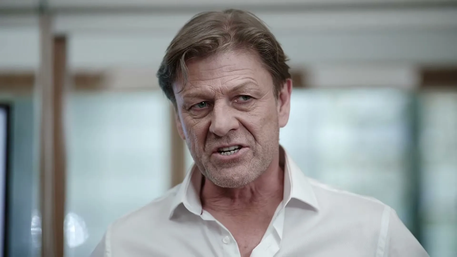 Sean Bean, a 60s+ male in a white shirt, speaks intently in an interior setting, with a serious expression.