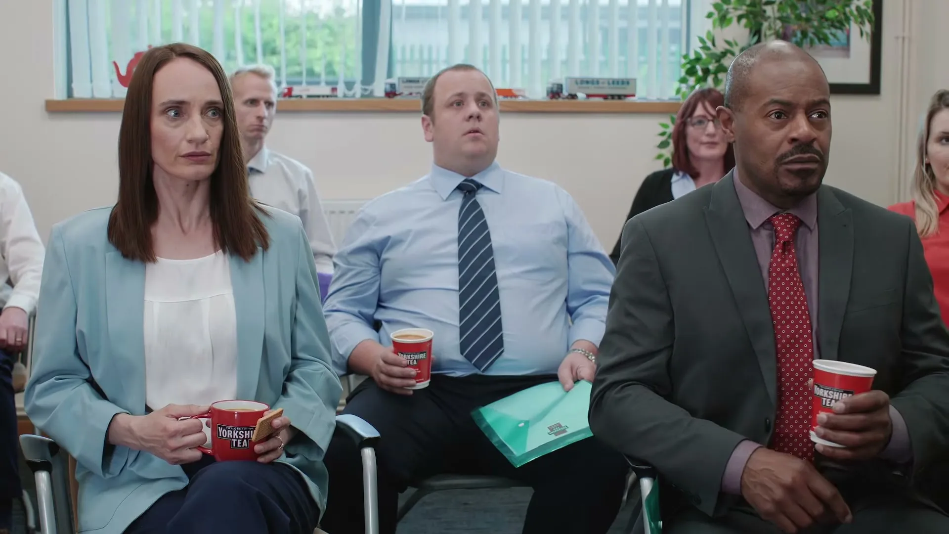 A diverse group of people, including a woman in a blue blazer and a man in a suit, sit in an office meeting room, holding Yorkshire Tea mugs and looking intently forward.