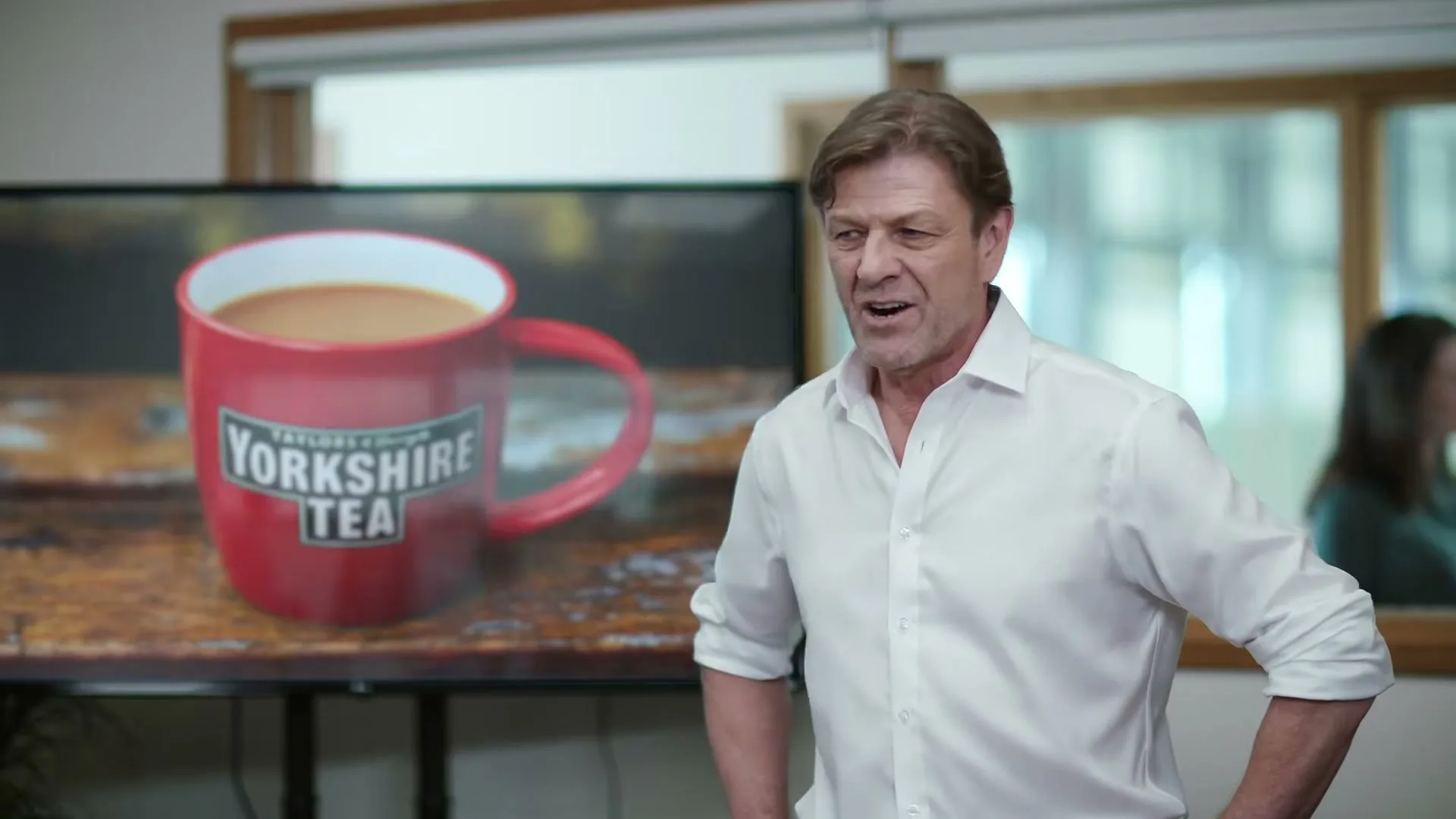 Sean Bean, a middle-aged man in a white shirt, stands next to a screen displaying a red mug of Yorkshire Tea, in an office.
