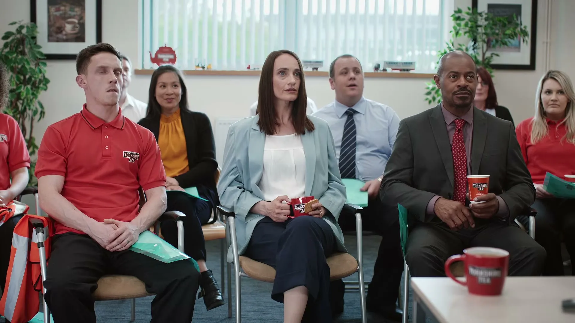 A diverse group of office workers sits in a meeting room, looking up and listening intently, some holding Yorkshire Tea mugs.
