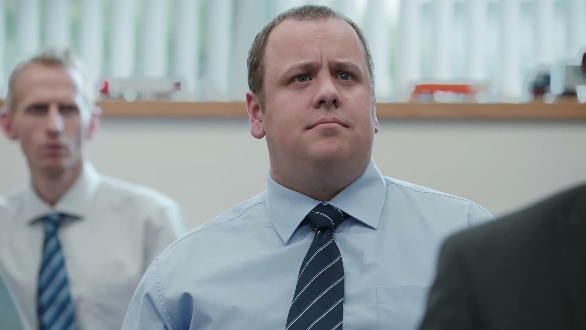 A man in a light blue shirt and striped tie looks up with a serious expression in an office setting.