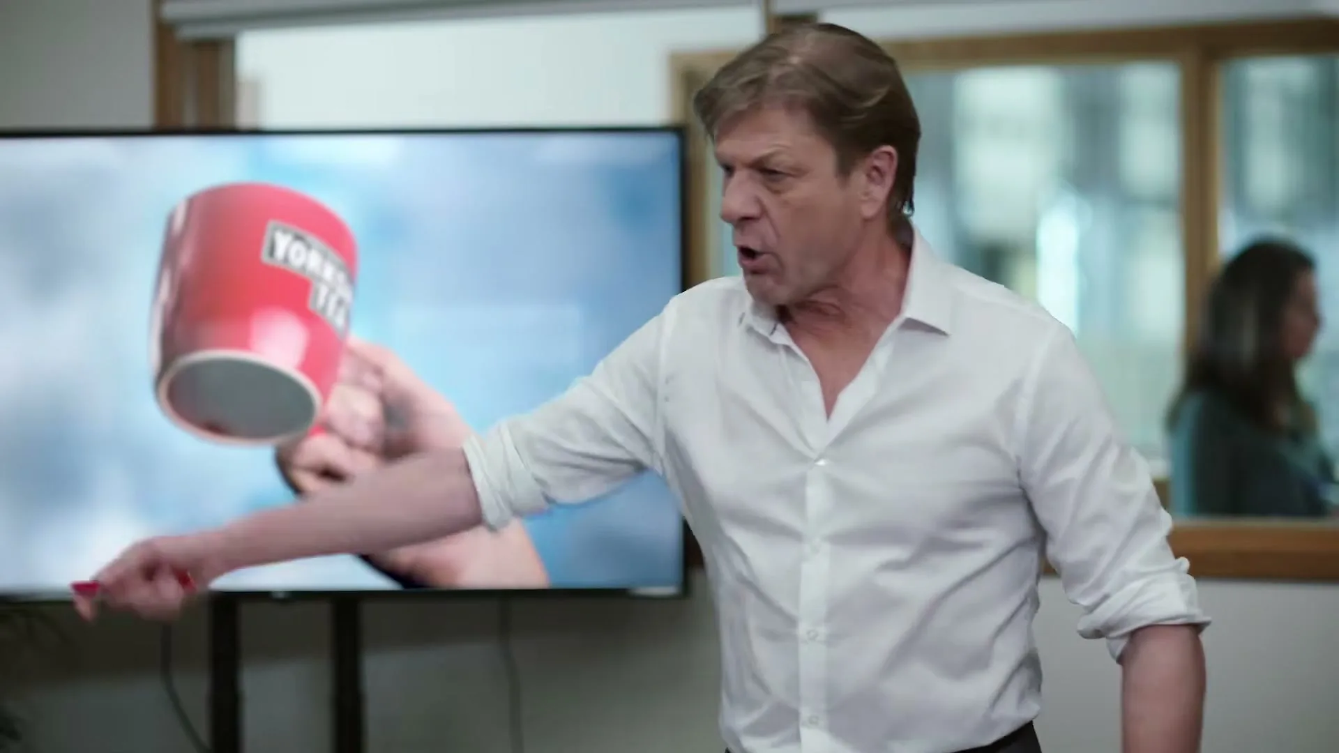 Sean Bean, a male in his 60s, wearing a white shirt, shouts and gestures with a pointer at a TV screen showing a red mug, in an office.