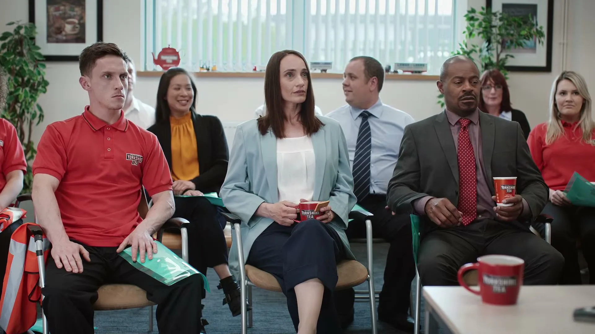 A diverse group of office workers sits in a meeting room, listening intently to an unseen speaker, some holding Yorkshire Tea cups.
