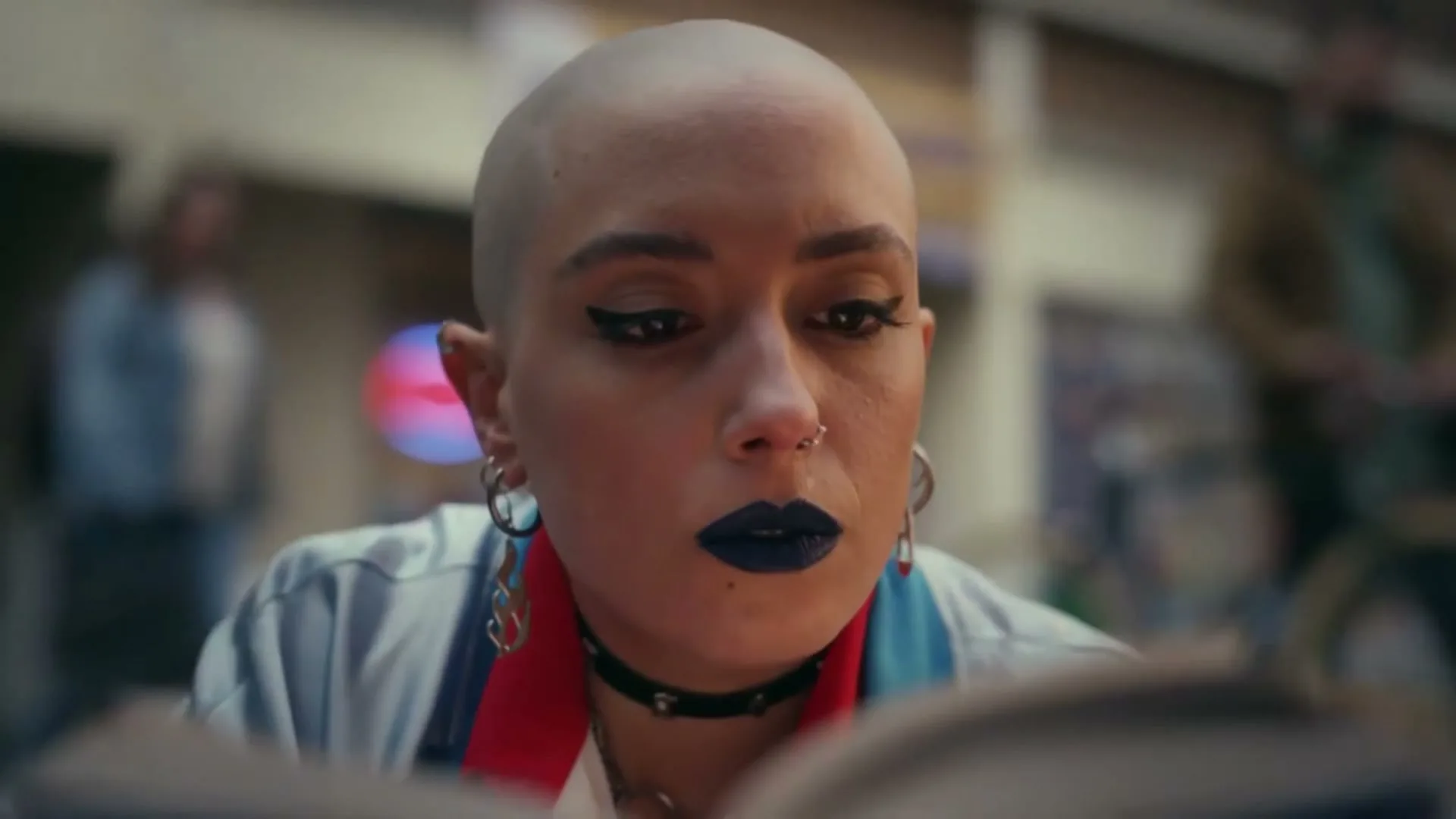 A young woman with a shaved head and dark lipstick reads a book intently, her face illuminated by the pages, in an outdoor setting.
