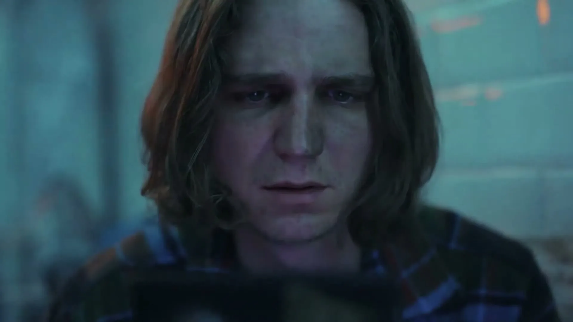 A man with long brown hair and a plaid shirt looks down with a concerned expression, illuminated by a blue light in a dark, possibly industrial, interior.