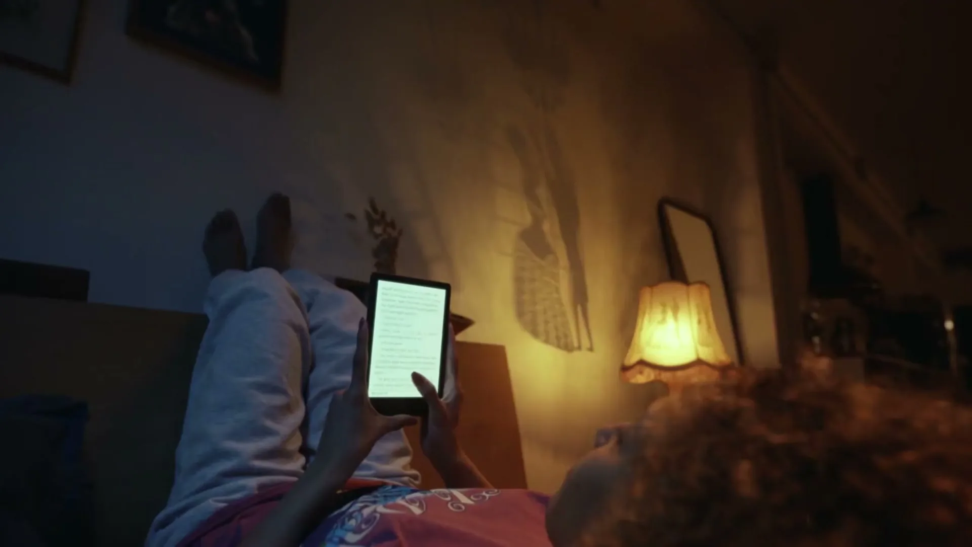A young woman with curly hair lies on a bed, holding an e-reader above her head, with her feet up against the wall in a dimly lit room.