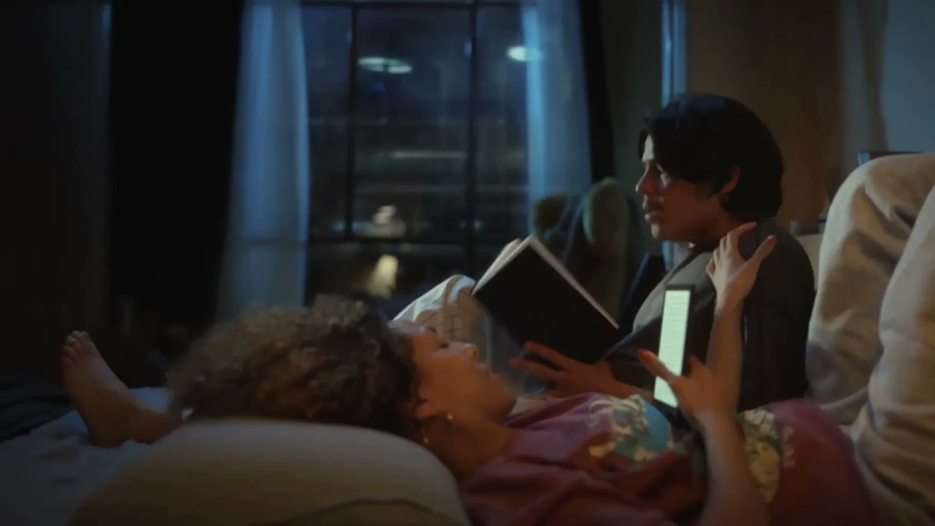 A young couple, a man and a woman, are lying in bed at night, reading books and talking to each other.