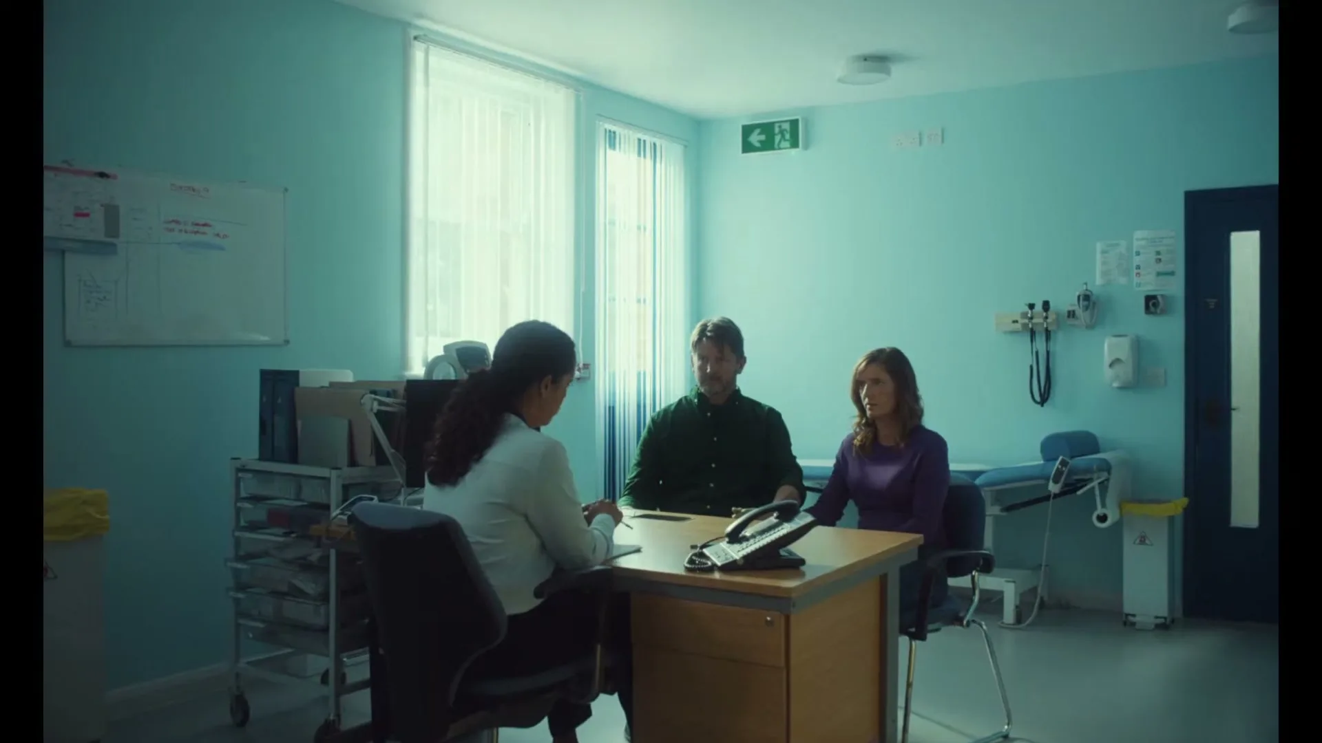 A female doctor consults with a male and female patient in a brightly lit hospital examination room, discussing an operation.