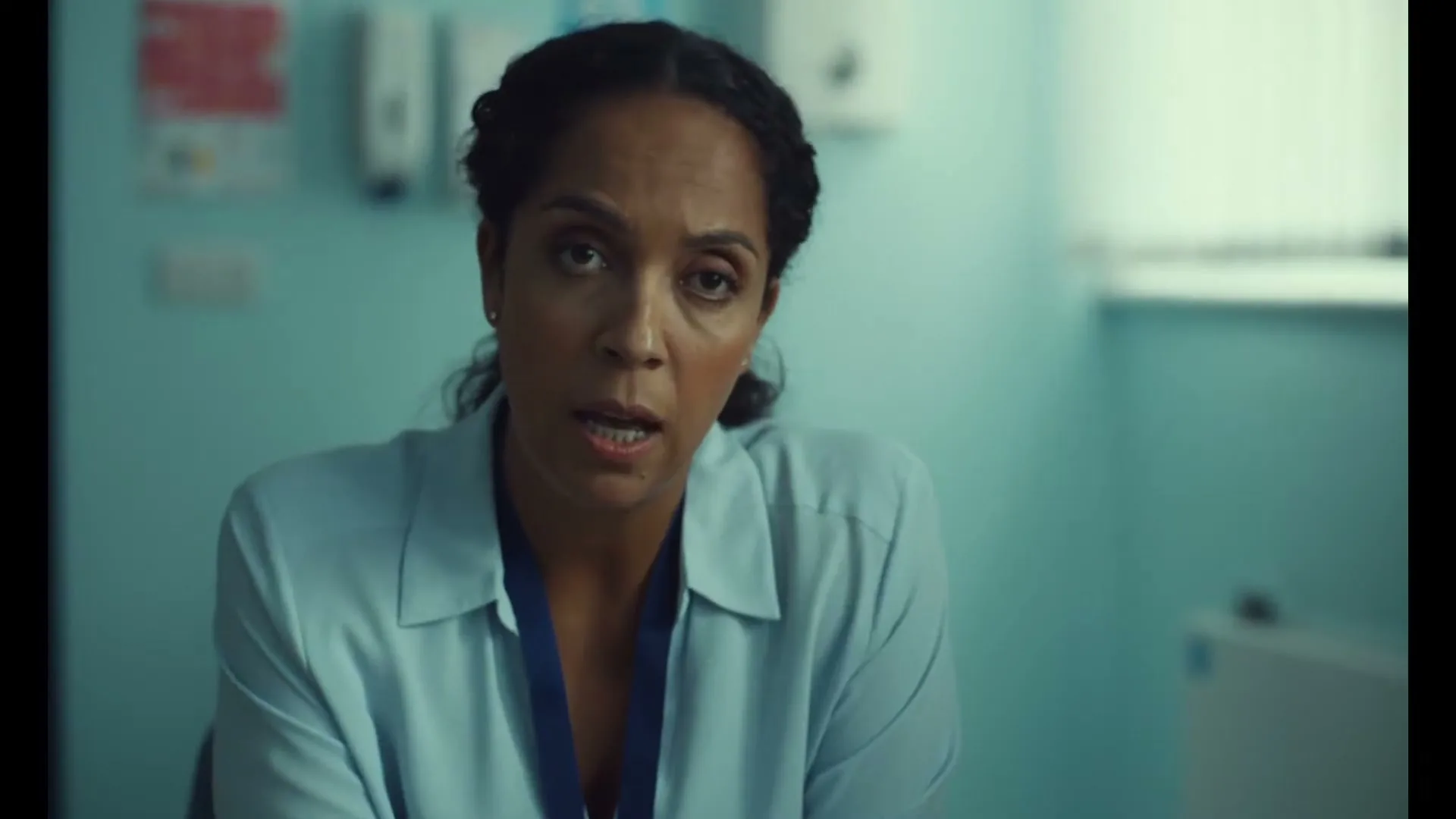 A female doctor with dark skin and braided hair, wearing a light blue uniform, speaks directly to the camera in a hospital room.