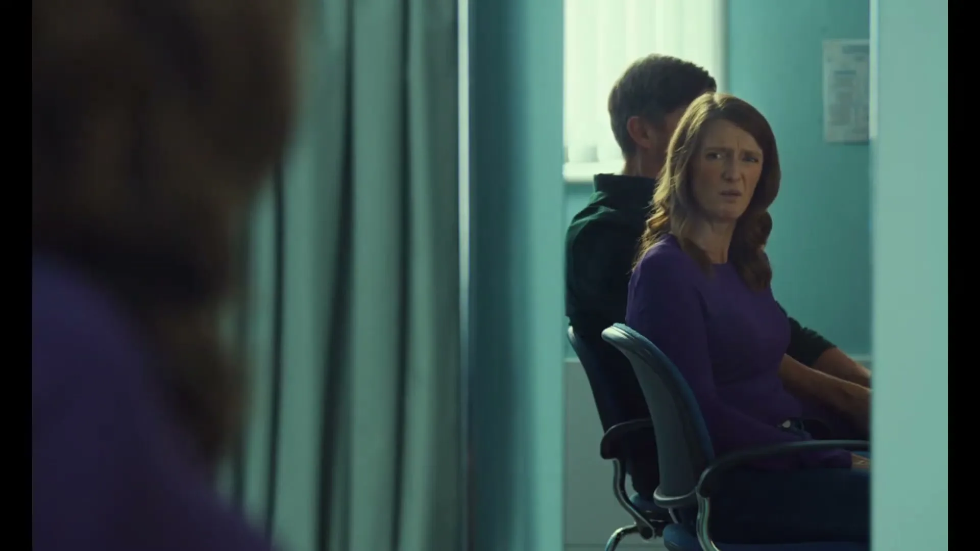 A woman with long brown hair and a purple top sits in a chair, looking worried, with a man behind her, in a doctor's office.