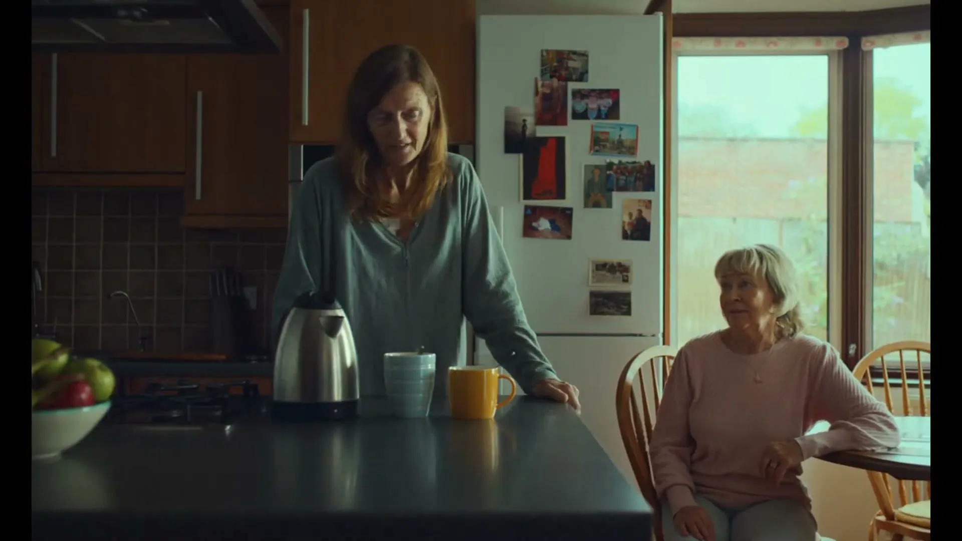 A woman in her 40s stands in a kitchen, looking down at two mugs on a counter, while an older woman sits at a table beside her.