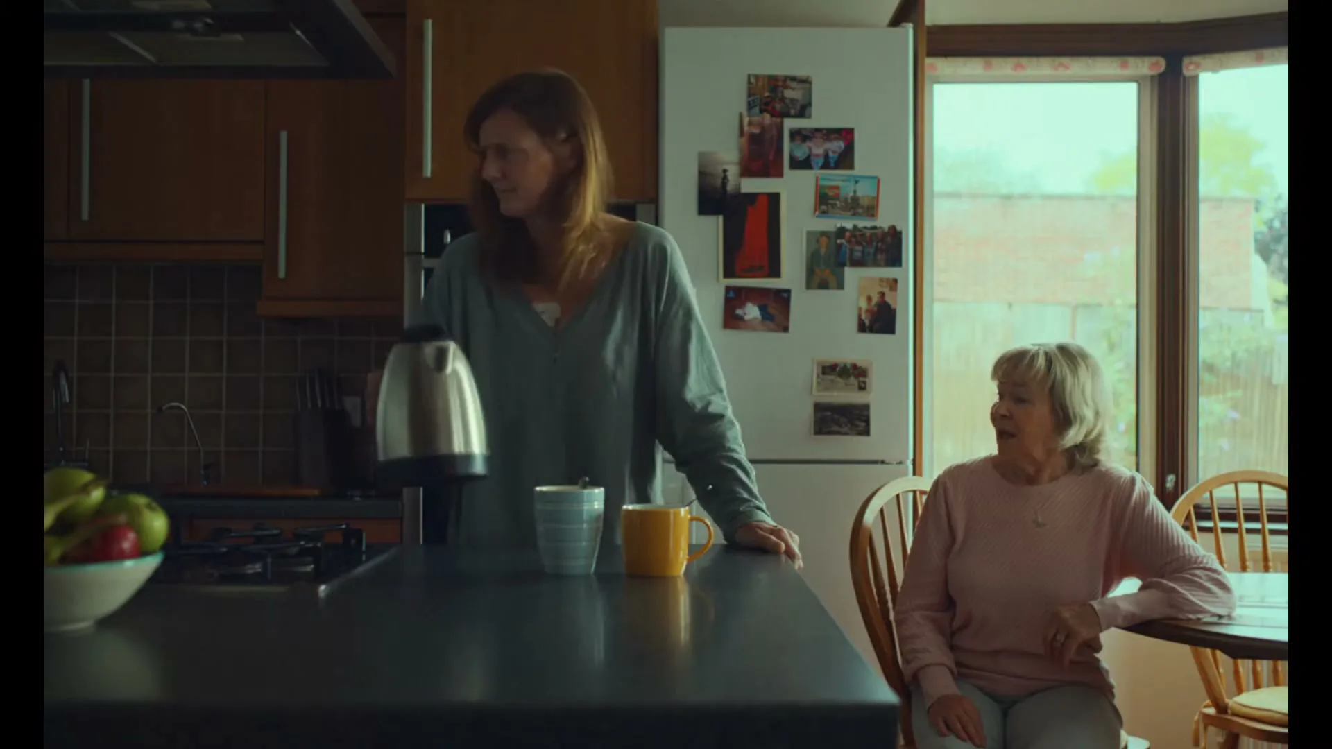 A woman in her 40s stands in a kitchen, holding a kettle, while an older woman sits at a table, looking at her.