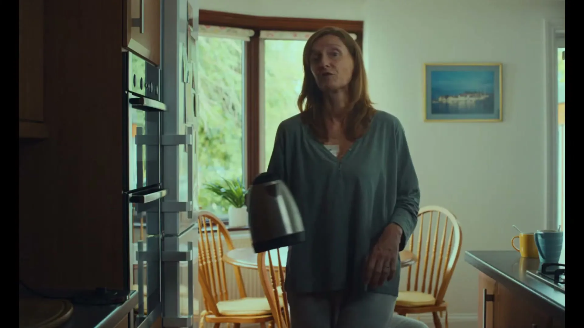 A woman in her 40s, wearing a grey top, holds a kettle and talks in a kitchen, with another woman visible in the background.