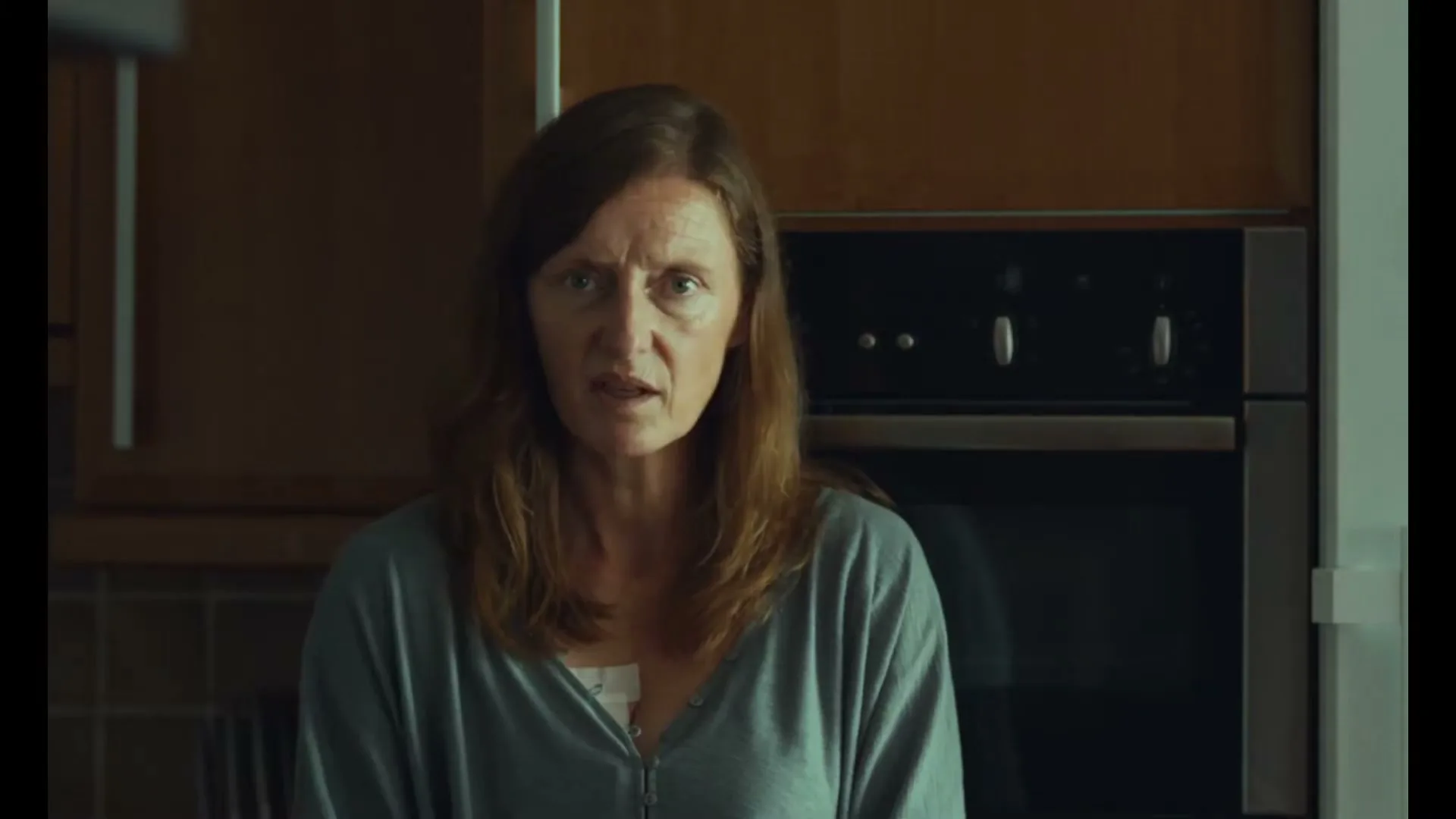 A woman with long brown hair and a grey top looks from smiling to serious in a kitchen.