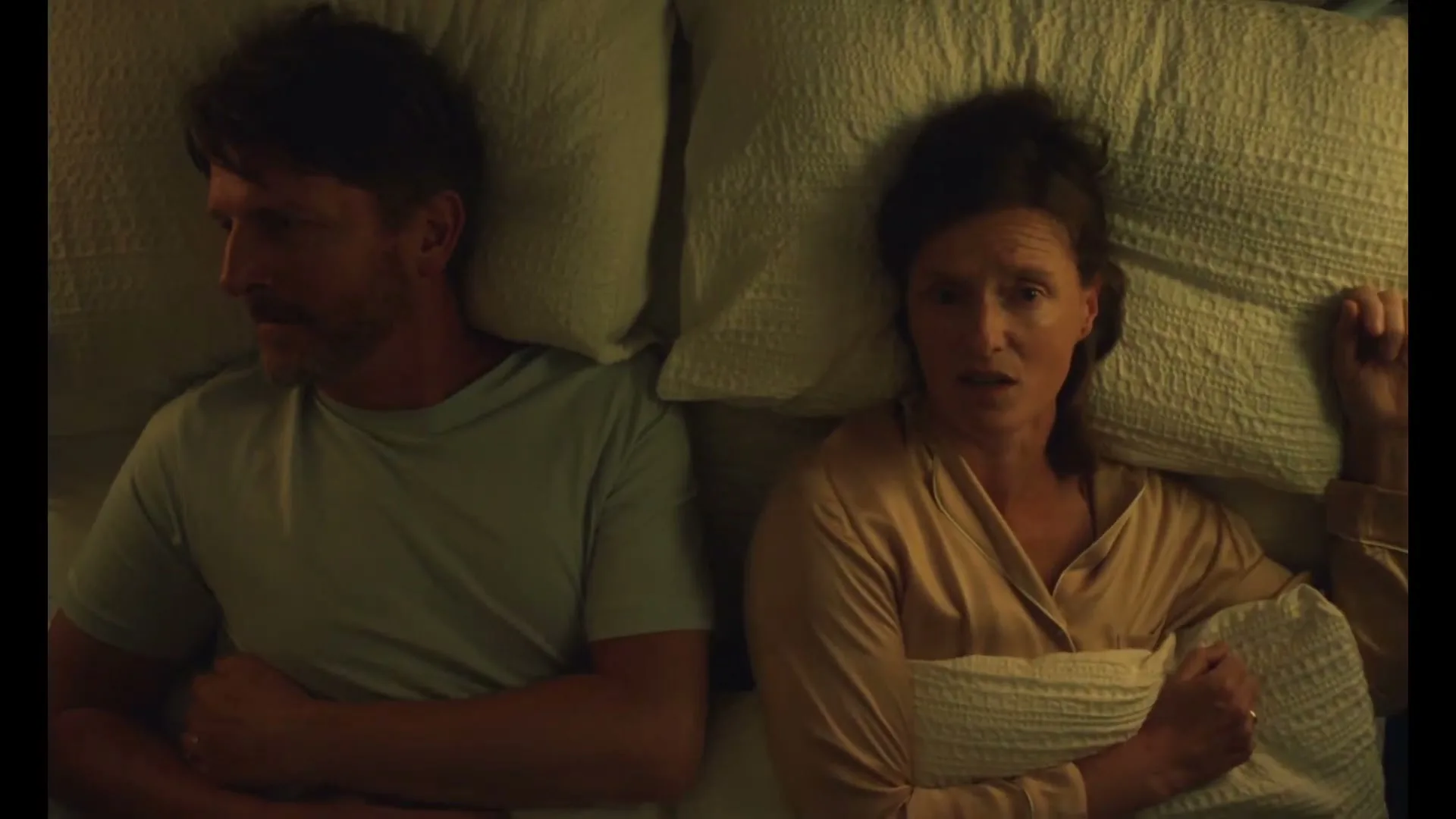 A middle-aged couple lies in bed, facing away from each other, looking distressed and thoughtful in a dimly lit bedroom.