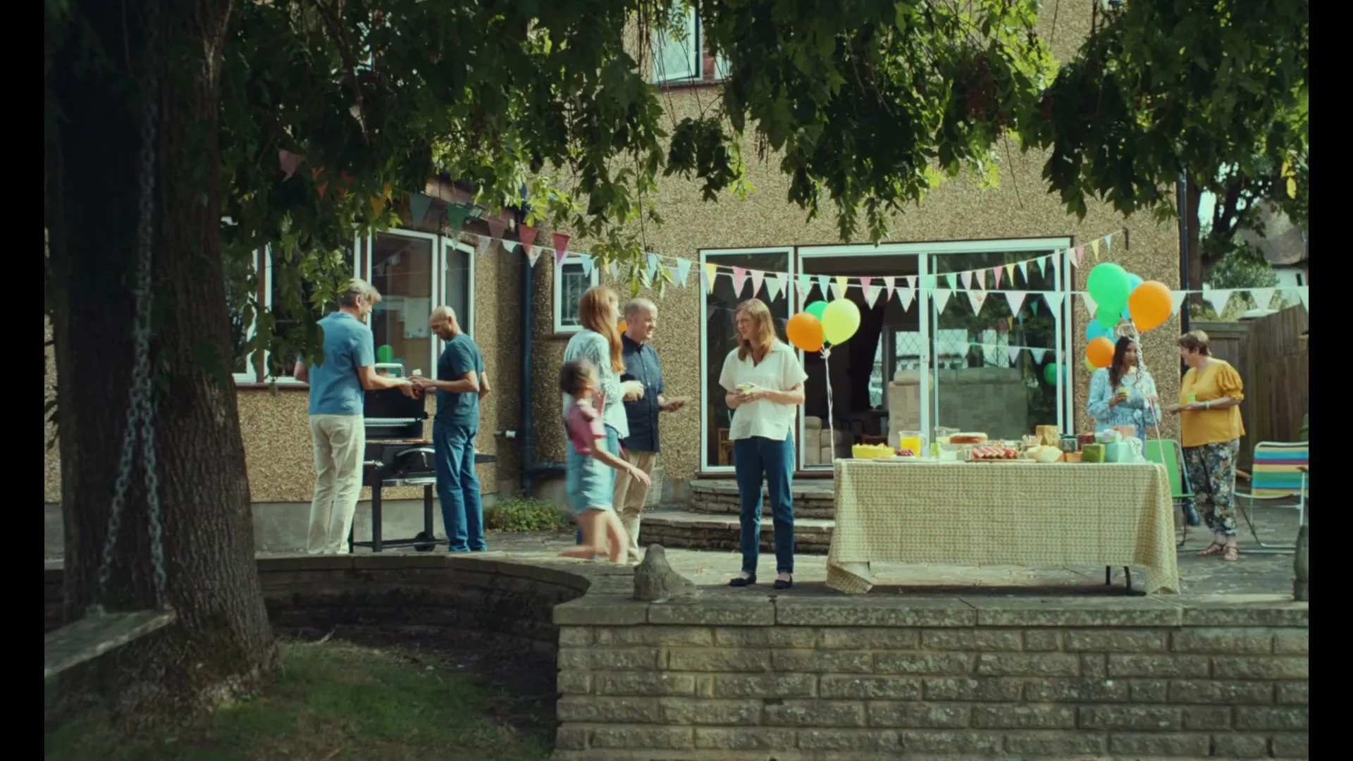 A diverse group of people of various ages gather in a suburban backyard for a festive outdoor party with balloons and food.