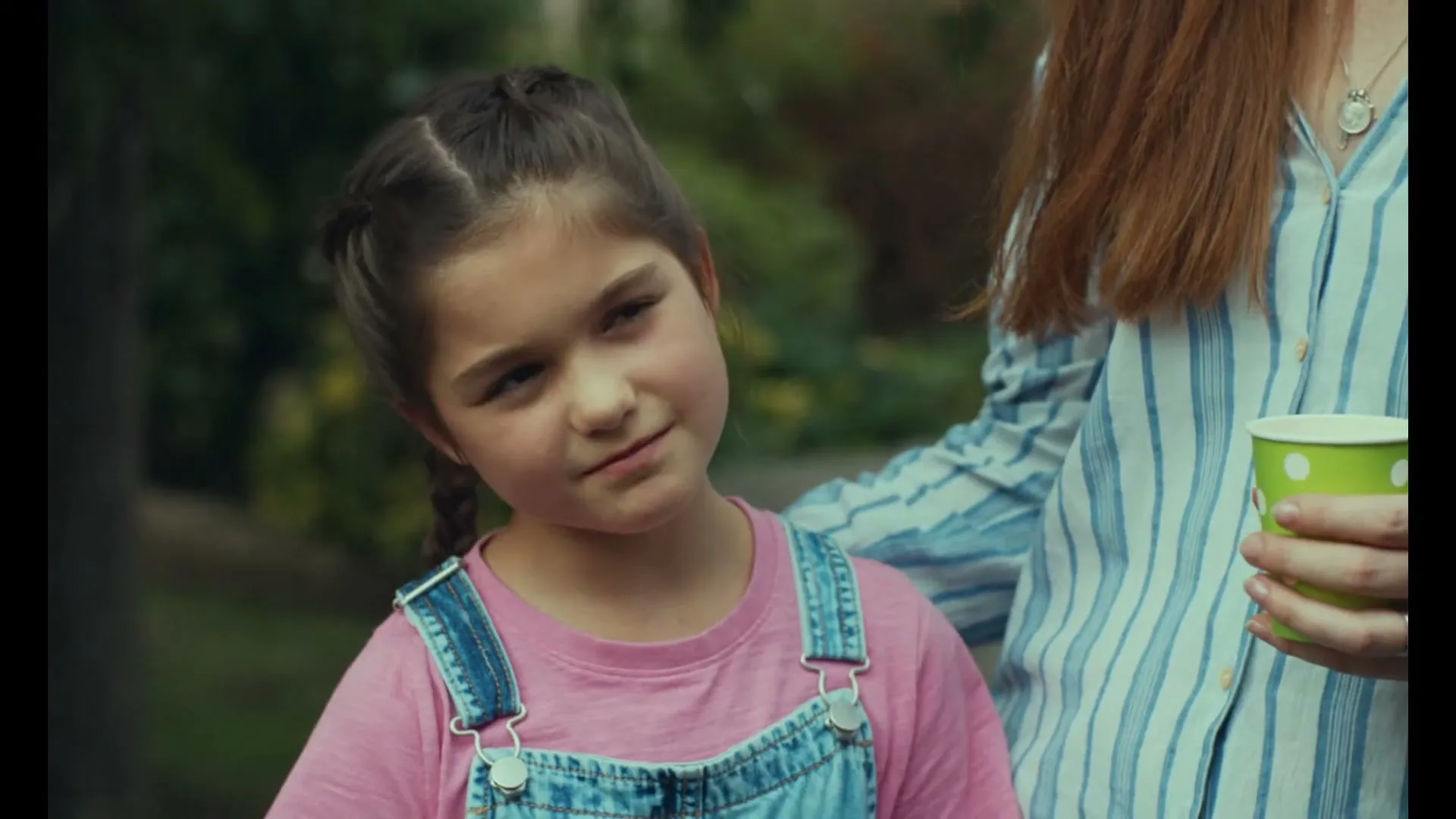 A young girl with braided hair and denim overalls looks up and smiles at an adult, who is partially visible holding a green cup in an outdoor setting.