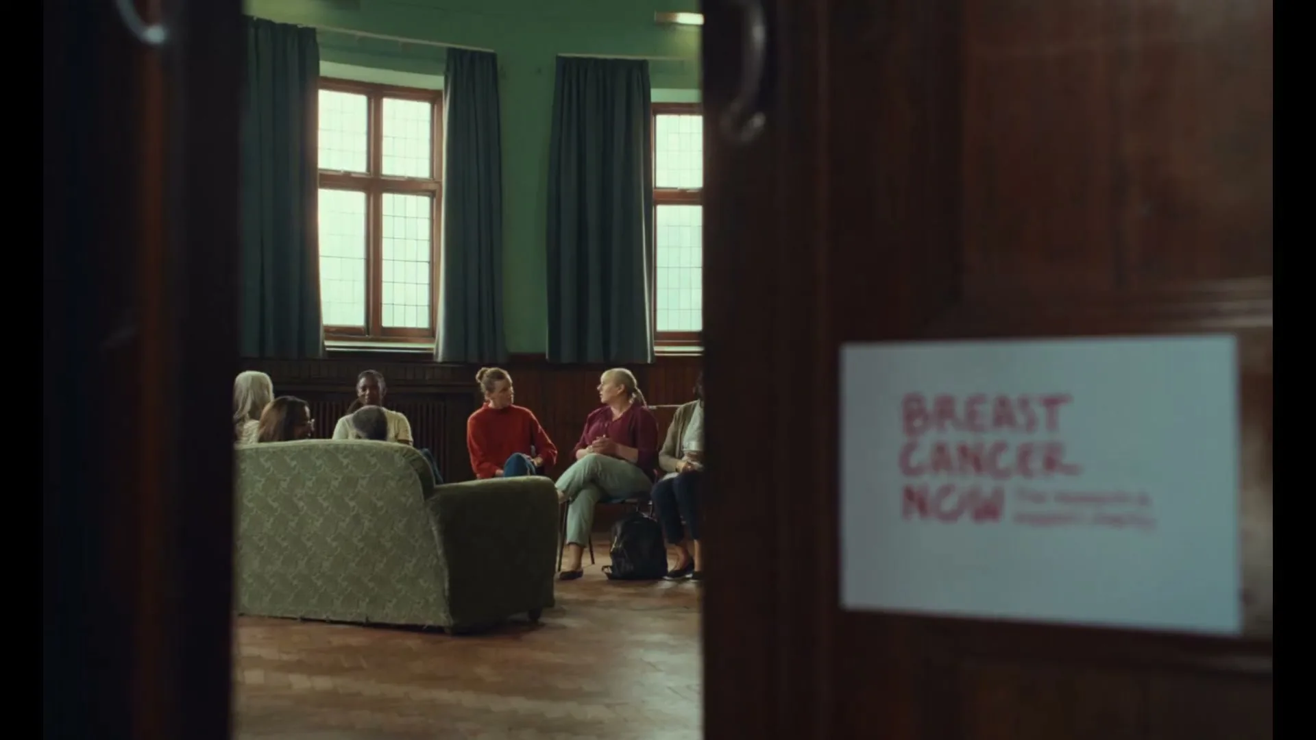 A diverse group of women sit in a circle, talking in a support group meeting, seen through an open doorway with a "Breast Cancer Now" sign.