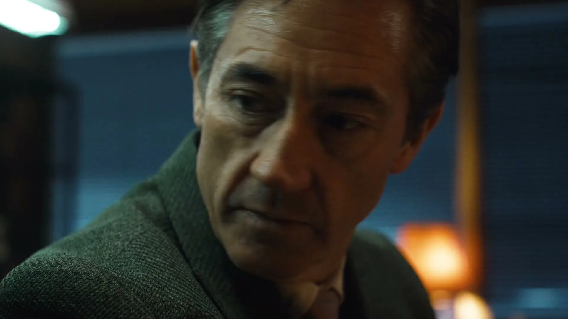 A man in his 40s, wearing a tweed jacket, looks down with a serious expression in a dimly lit interior.