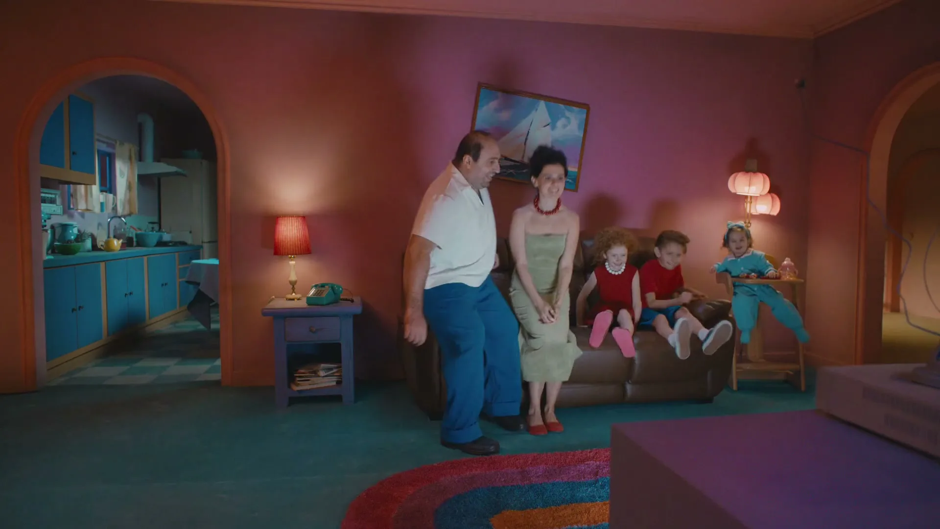 A family of five, dressed in colorful clothes, runs into a living room and sits on a brown couch, in a house with purple walls and blue kitchen cabinets.