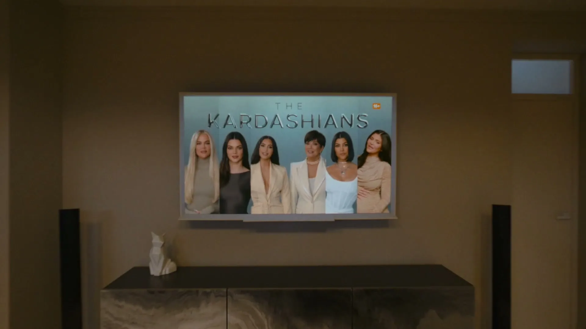 A television screen displays "The Kardashians" with five women, as the camera pulls back to reveal a minimalist living room with two people walking past.