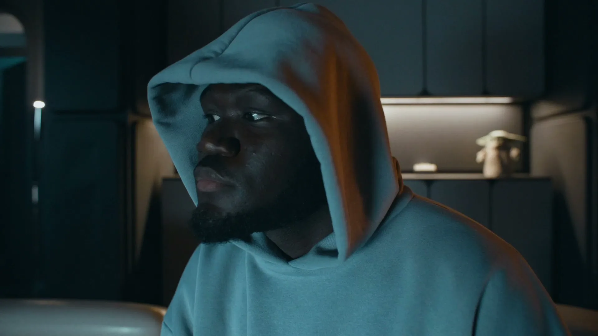 A young Black man in a blue hoodie looks up and then down, illuminated by a screen in a dark, modern interior.