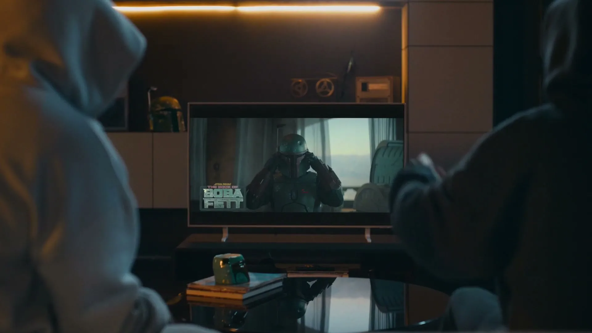 Two people in hoodies watch "The Book of Boba Fett" on a TV in a dimly lit living room, with one person pointing at the screen.