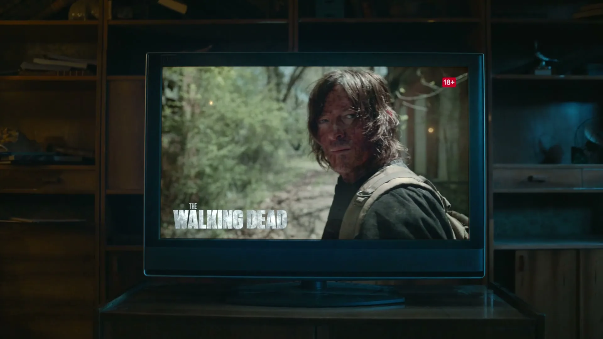 A man with long hair and a dirty face looks over his shoulder on a TV screen, showing "The Walking Dead" in a dimly lit room.