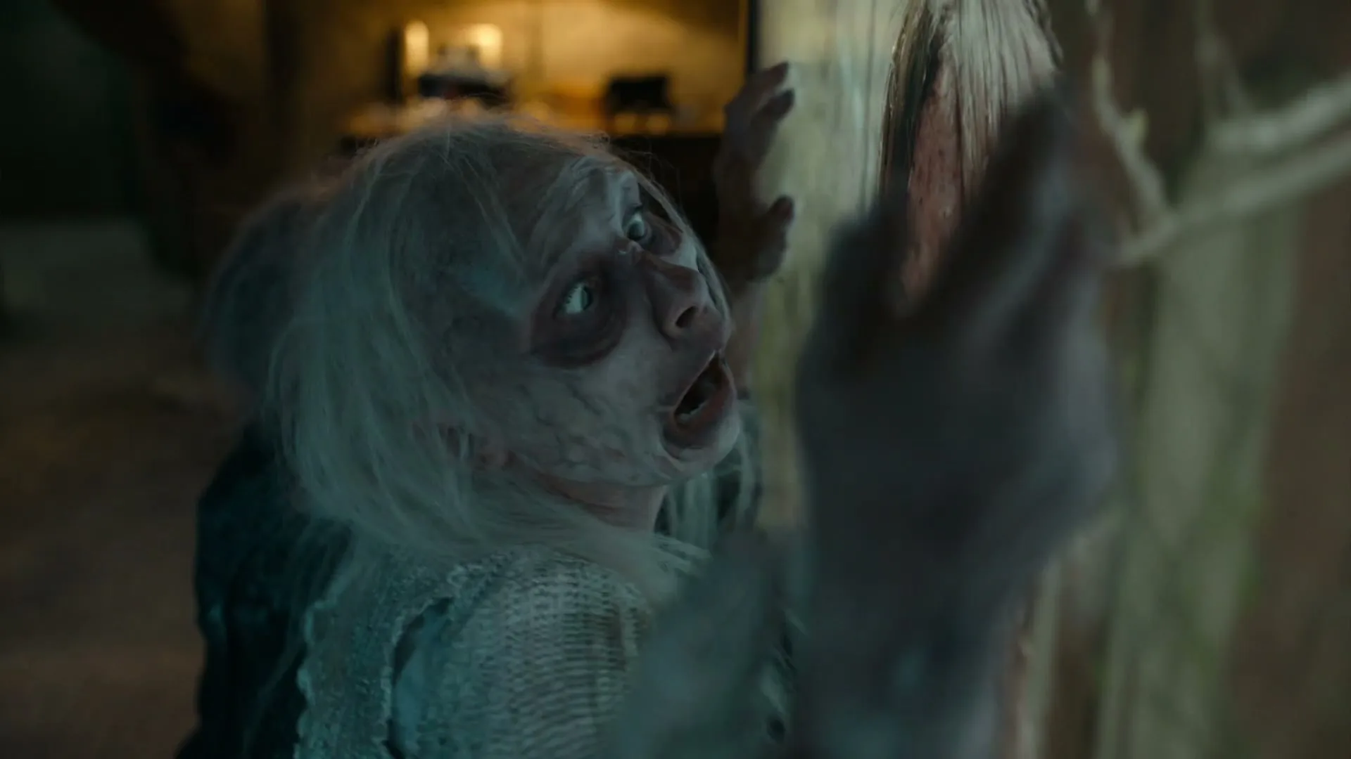A woman with white hair and zombie-like makeup looks up in fear, pressing against a wall in a dimly lit, spooky interior.