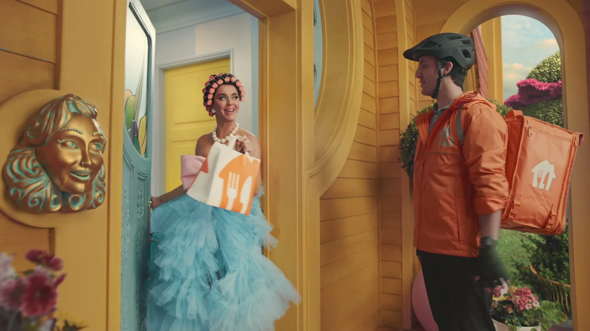 Katy Perry, dressed in a blue ruffled dress and pink bow, opens her front door to receive a food delivery from a male delivery driver in an orange jacket.