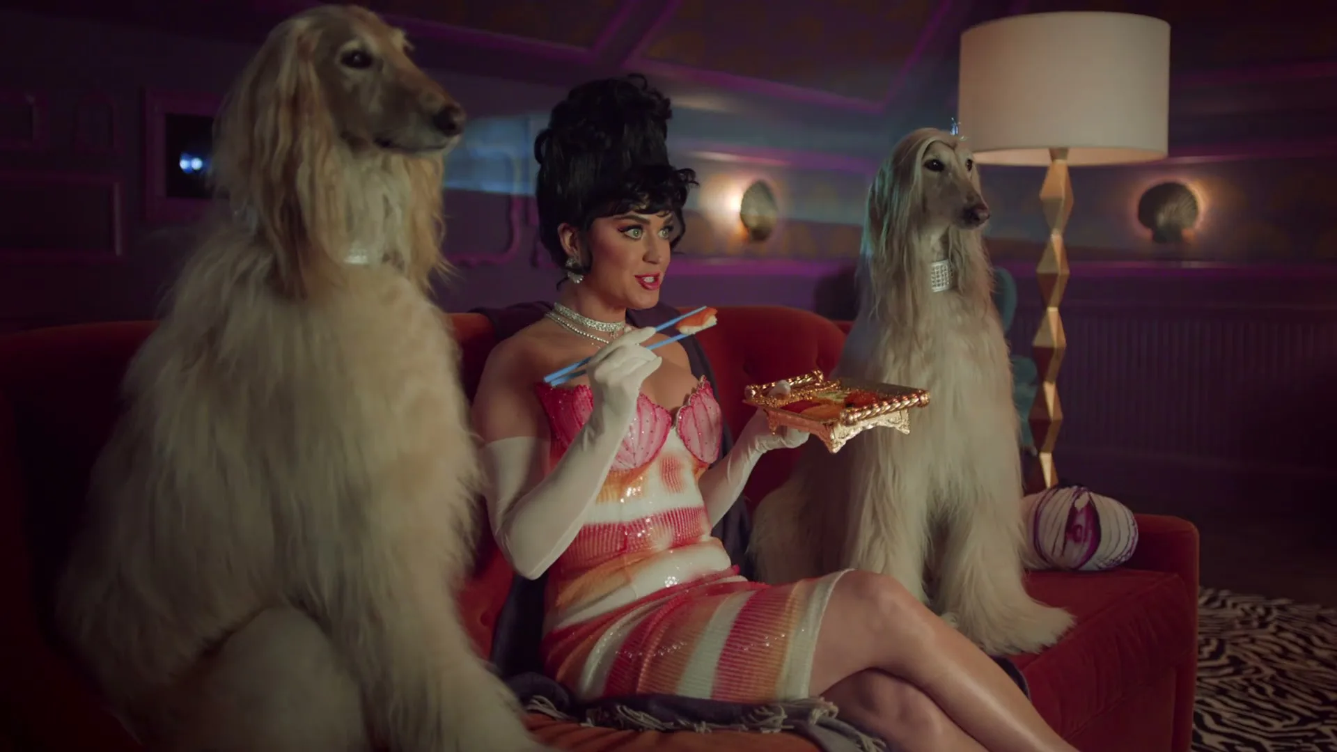 Katy Perry, dressed in a colorful sequined dress and white gloves, sits on a red couch between two Afghan Hounds, eating sushi in a dimly lit living room.
