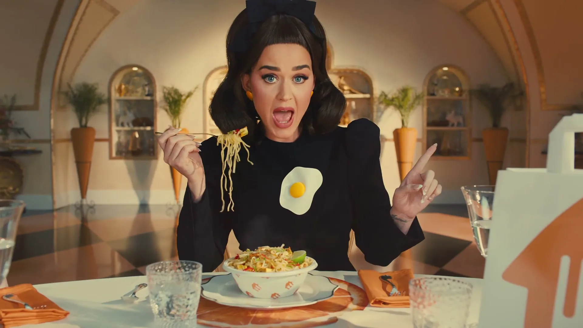 Katy Perry, dressed in various outfits, sings and dances in a surreal, dollhouse-like setting, interacting with different food items and a piano.