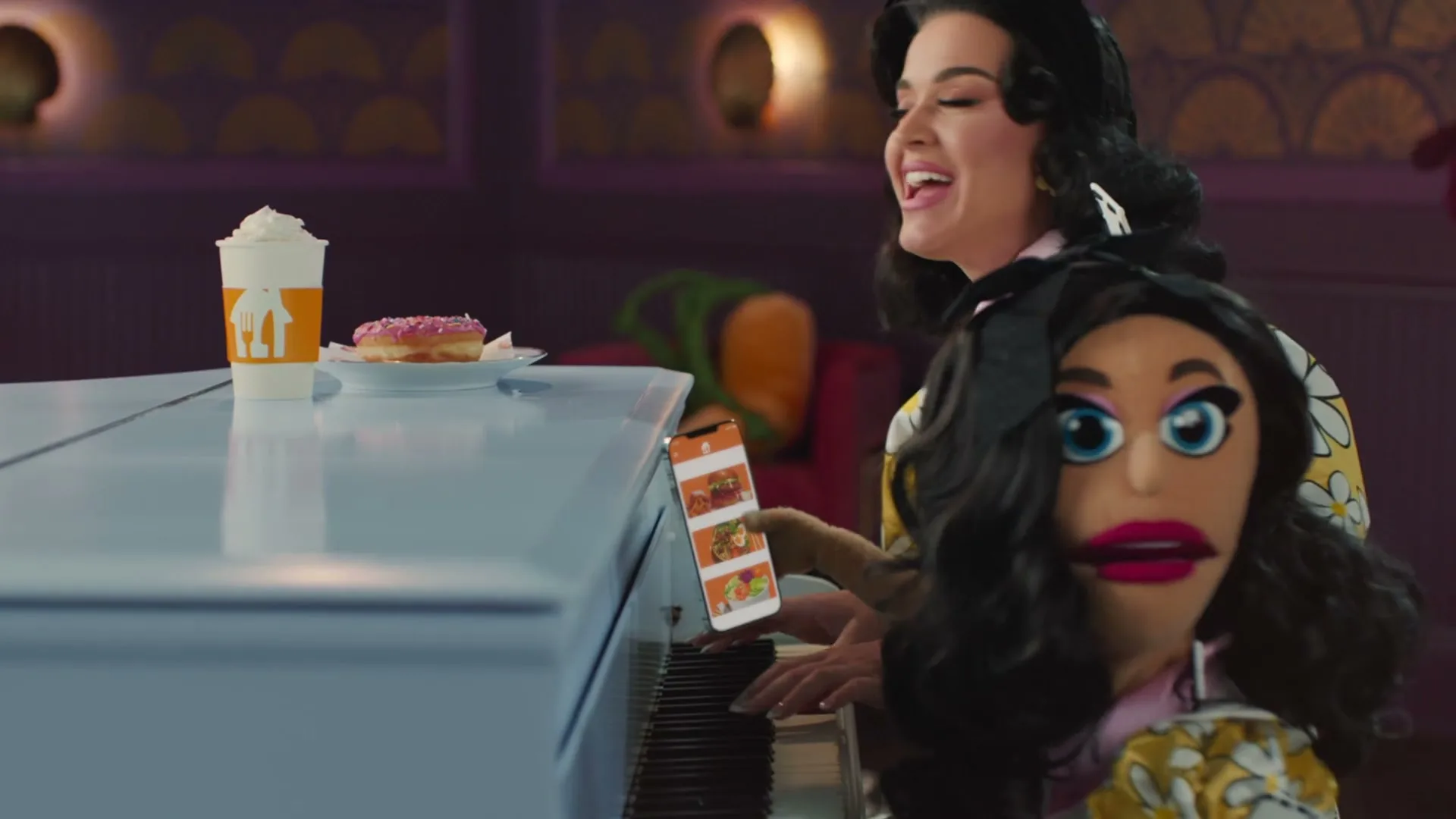 Katy Perry and a puppet version of herself play a white piano in a whimsical, purple-lit room, with a drink and donut on the piano.