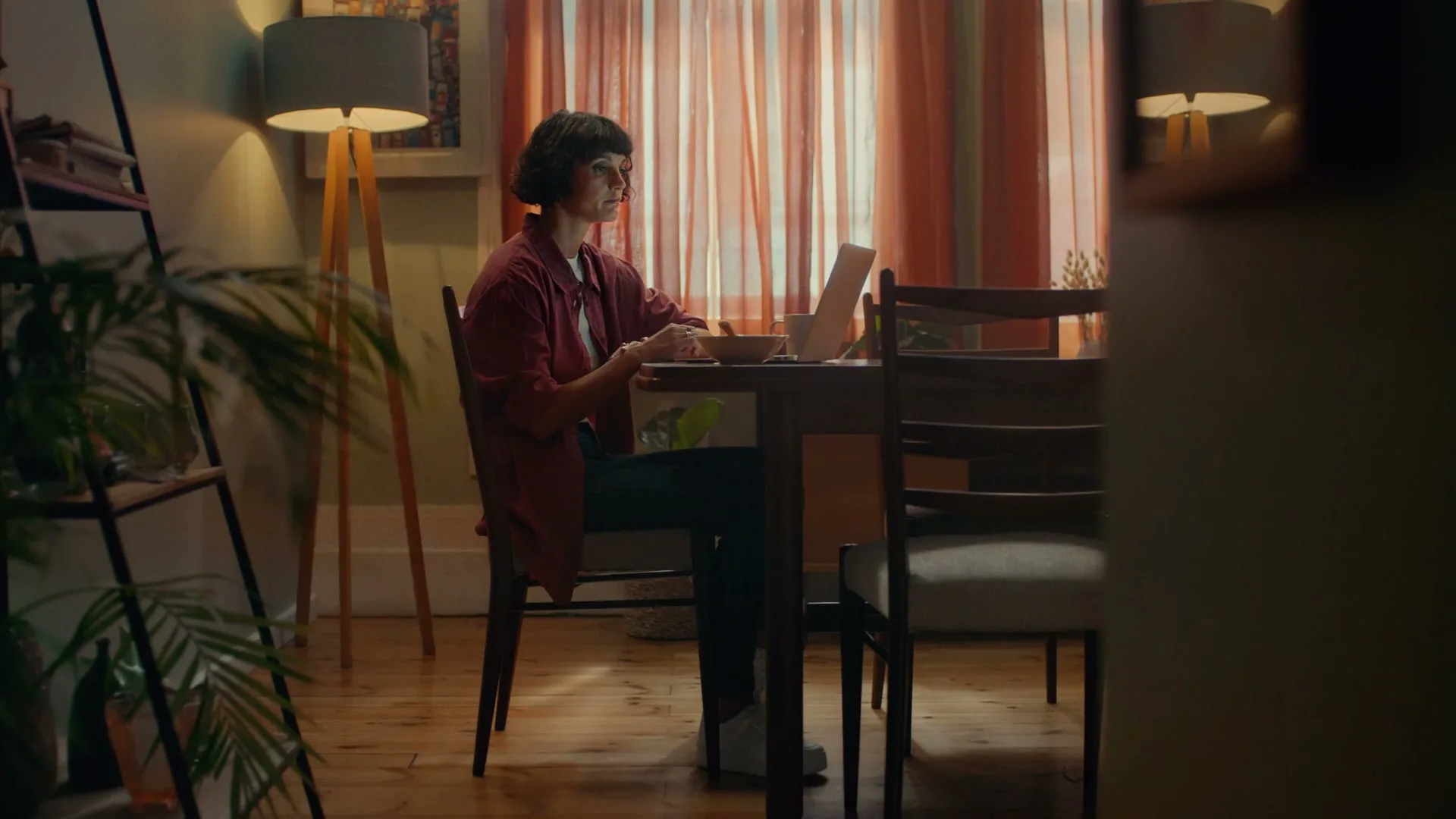 A young woman with short dark hair sits at a dining table, looking at a laptop while eating from a bowl in a cozy, dimly lit room.
