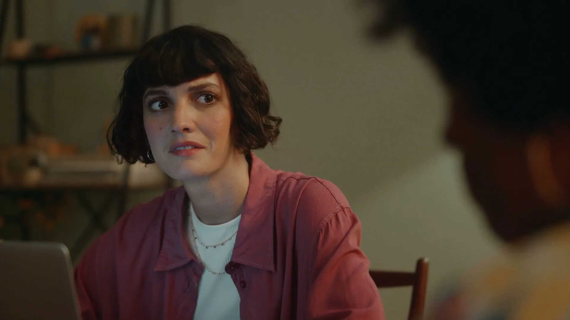A woman with short dark hair and a pink shirt looks intently at another person across from her in a dimly lit room.
