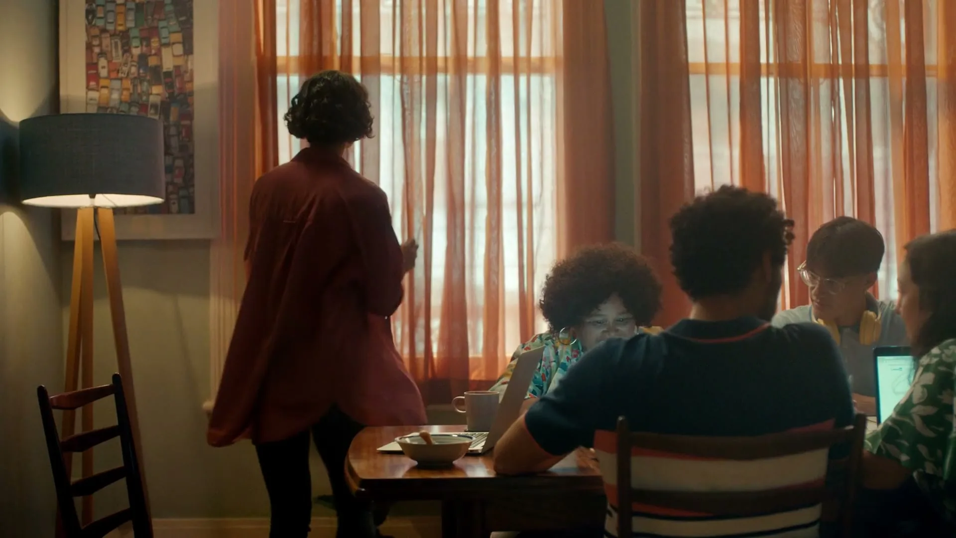 A diverse group of young adults works on laptops around a dining table, while a woman in a red shirt walks past them in a sunlit room.