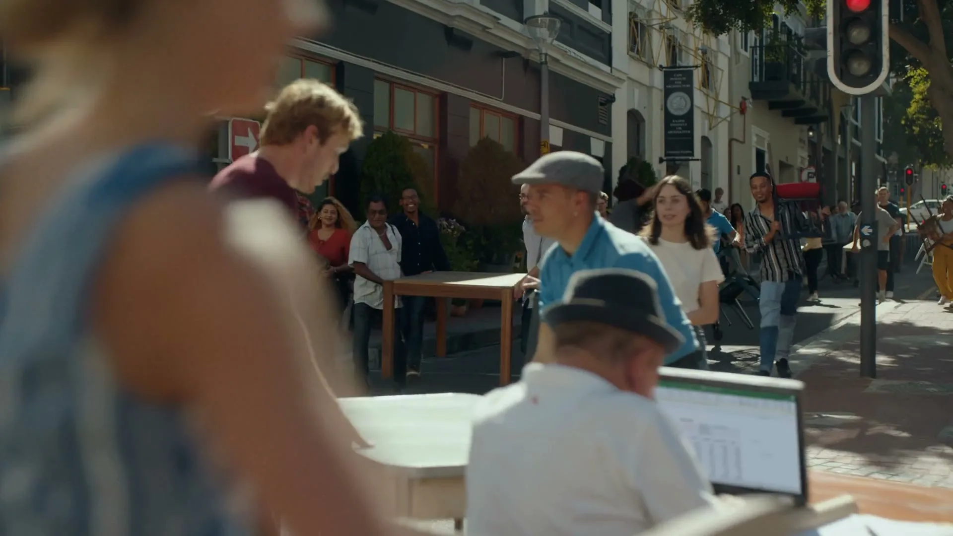 A diverse group of people, including a young man in a maroon shirt, are moving furniture and other items down a busy city street during the day.