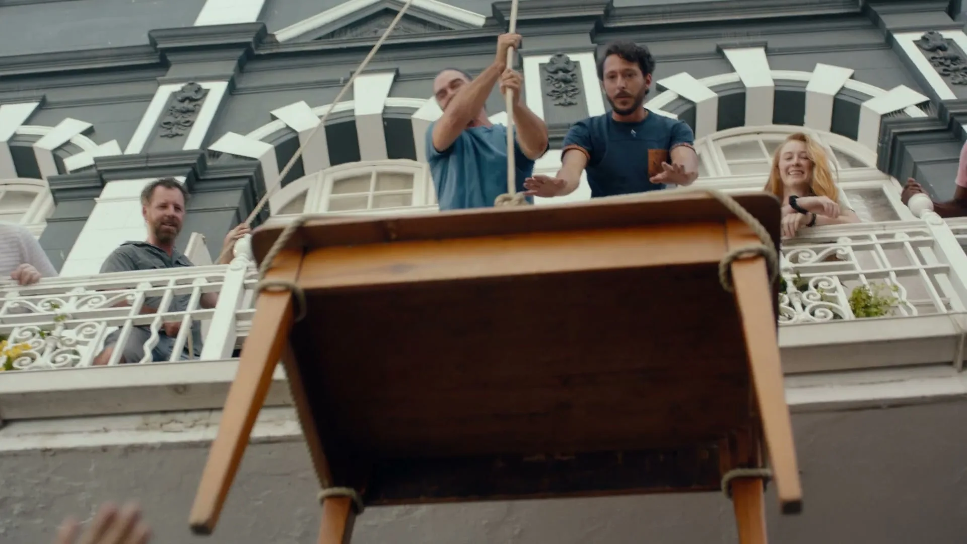 A group of diverse people on balconies watch as a wooden table is lowered by ropes from an upper floor of a building.
