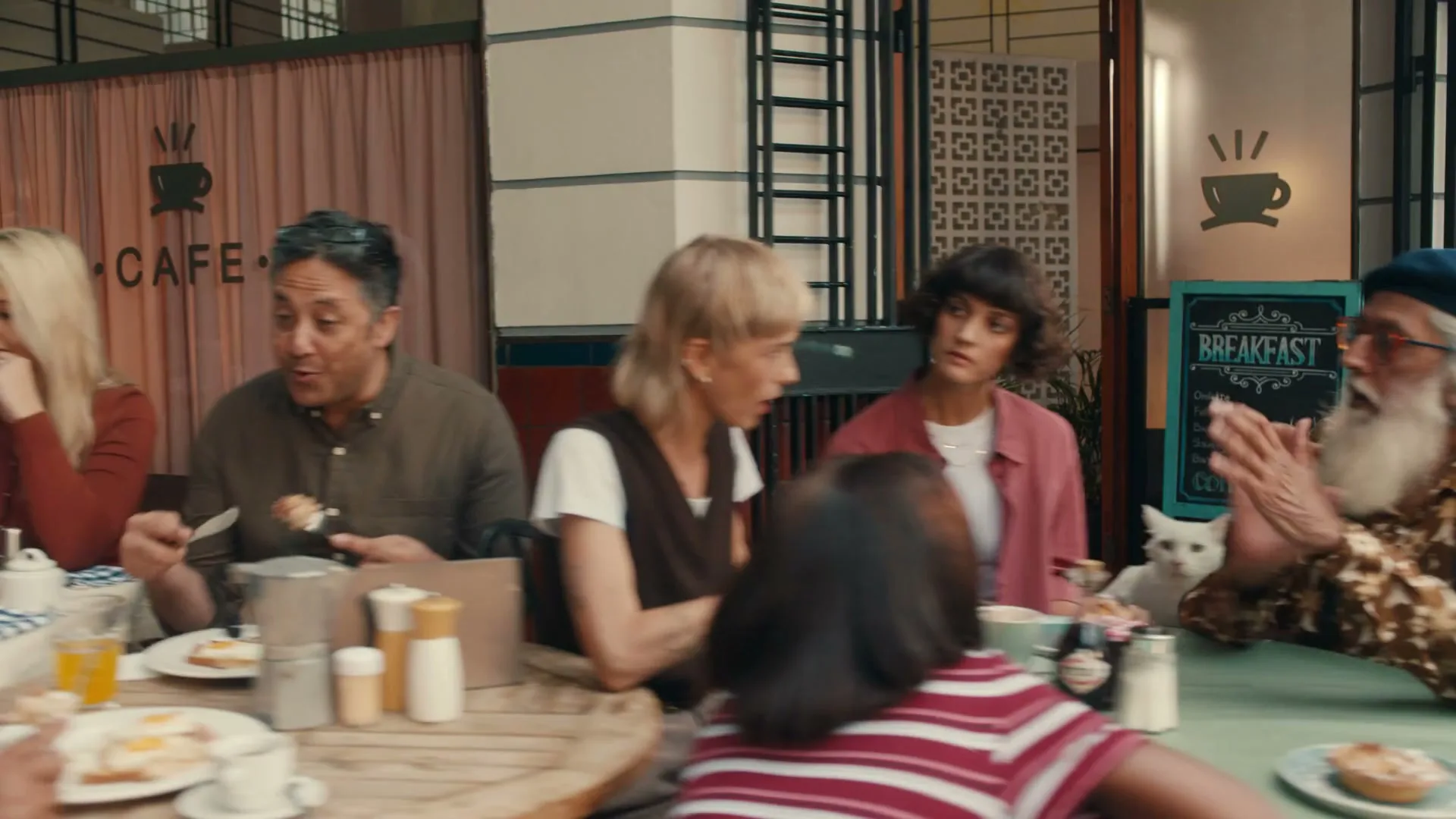 A diverse group of people of various ages are seated around tables, eating and conversing in a bustling cafe.