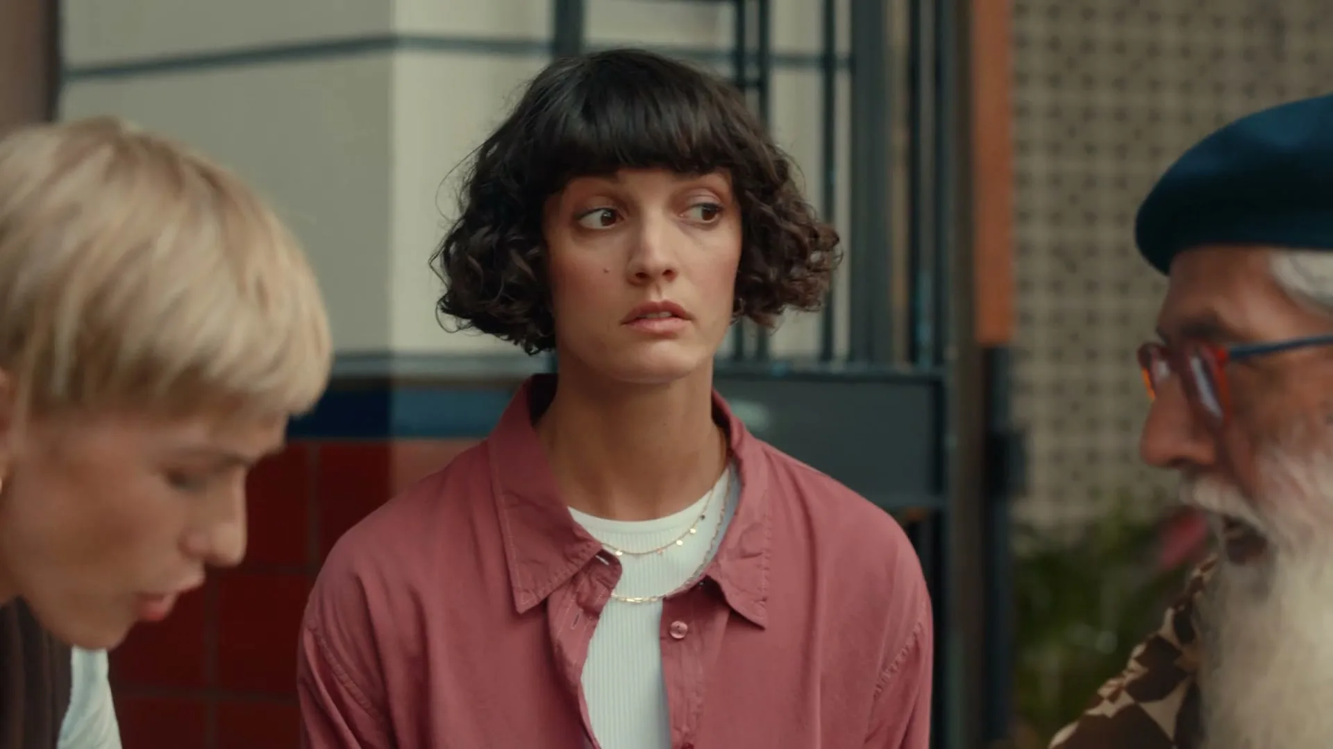 A young woman with short, dark curly hair and a pink shirt looks surprised while sitting between two other people in an outdoor setting.