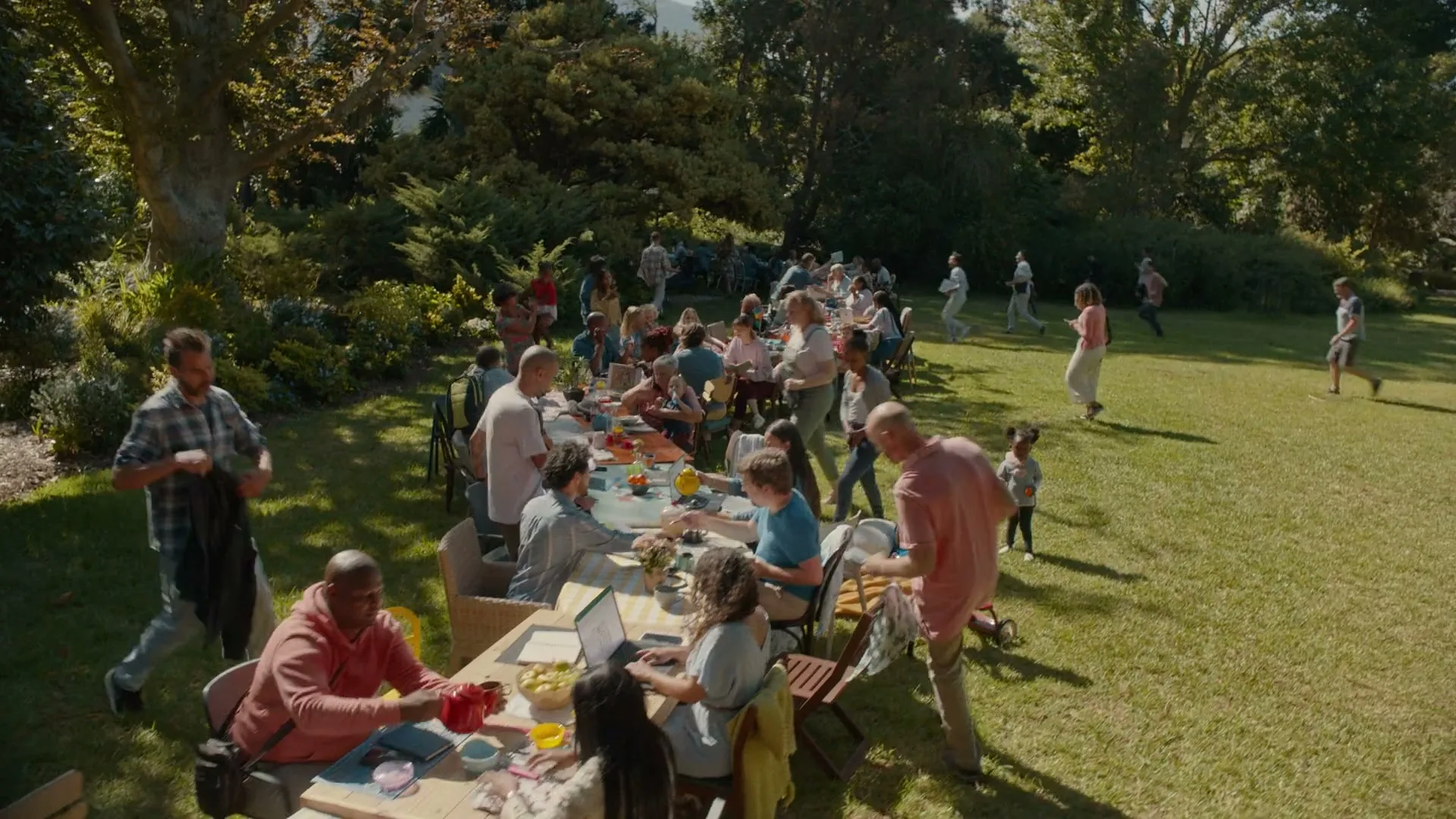 A diverse group of people of all ages gather for a large outdoor picnic on a sunny day, with some eating and others playing in the background.