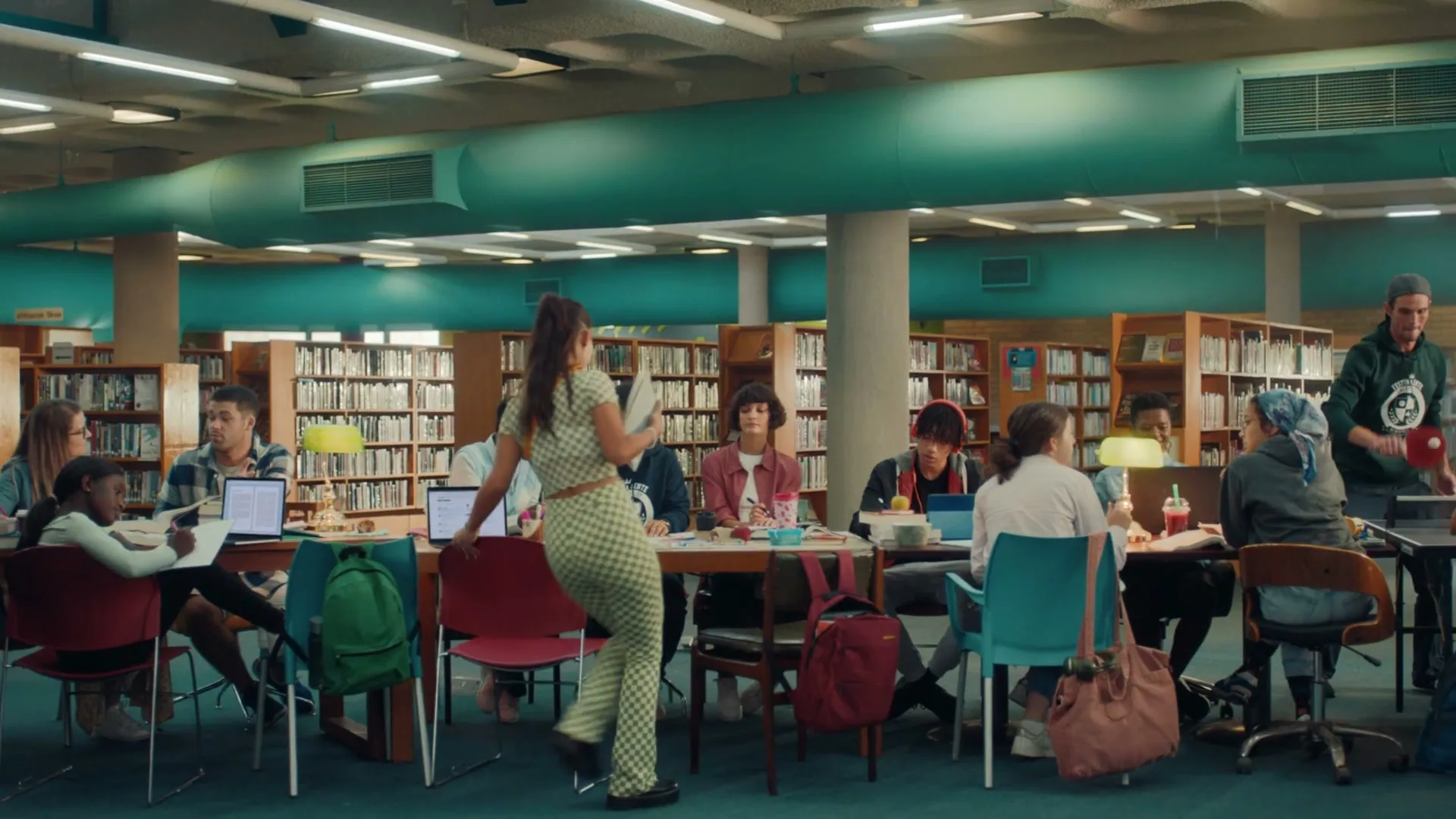 A diverse group of college students study and socialize at tables in a library, while one woman walks past in the foreground.