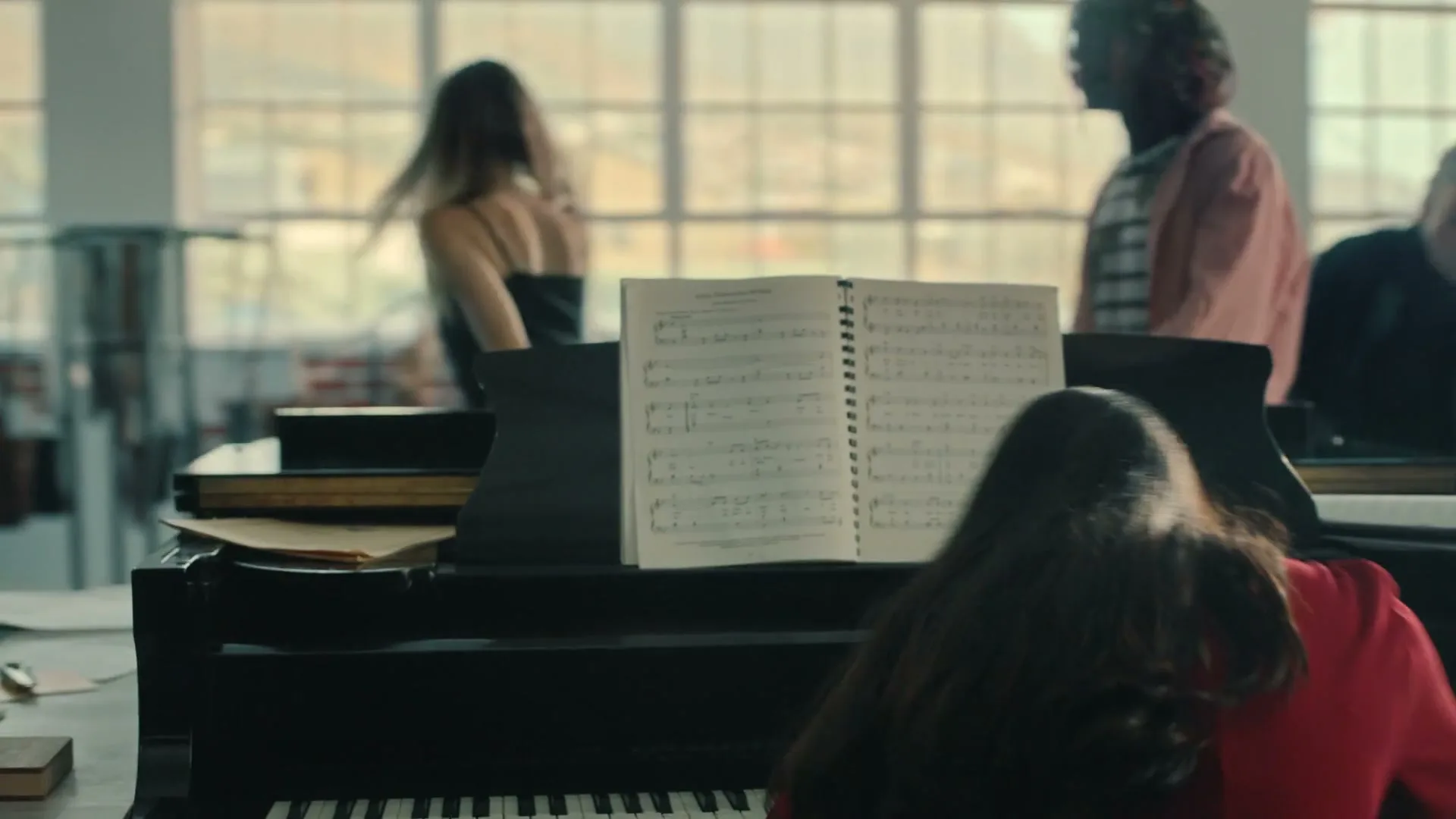A young woman with long dark hair plays a piano with sheet music in front of her, while other people move around in the background of a bright, airy room.