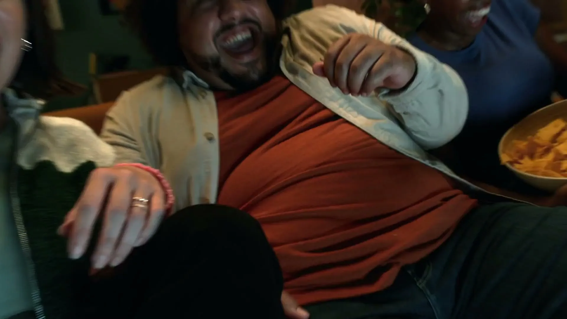 Three friends on a couch, two women and one man, scream in terror while watching TV in a living room at night.